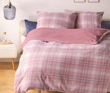 Ein Bett mit rosa karierter Bettdecke und Kissen sowie einer einfarbigen rosa Decke. Ein dunkelblaues Kissen liegt hinter den karierten Kissen. Links steht ein weißer Hocker mit einer Glasflasche und zwei Gläsern unter einer gerippten grauen Pendelleuchte. Zwei gerahmte Strichzeichnungen hängen an der Wand über dem Bett.