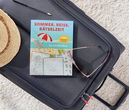 Auf einem schwarzen Koffer liegen ein Buch mit dem Titel Sommer, Reise, Rätselzeit, eine Sonnenbrille, ein Bleistift und ein Strohhut.