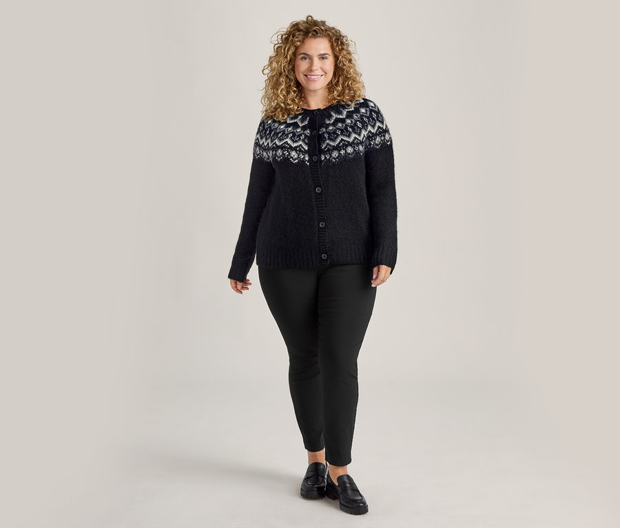 Frau mit lockigem Haar, die eine schwarze Strickjacke mit weißem Muster und eine schwarze Stretchhose trägt.