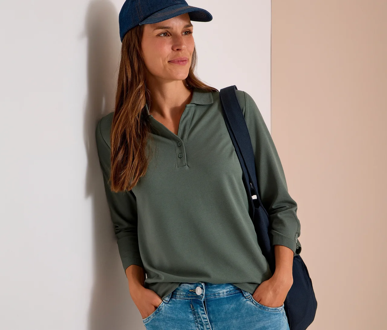Frau in grünem CECIL 3/4-Poloshirt, Jeans und Kappe, lehnt an einer Wand, Hände in den Taschen.