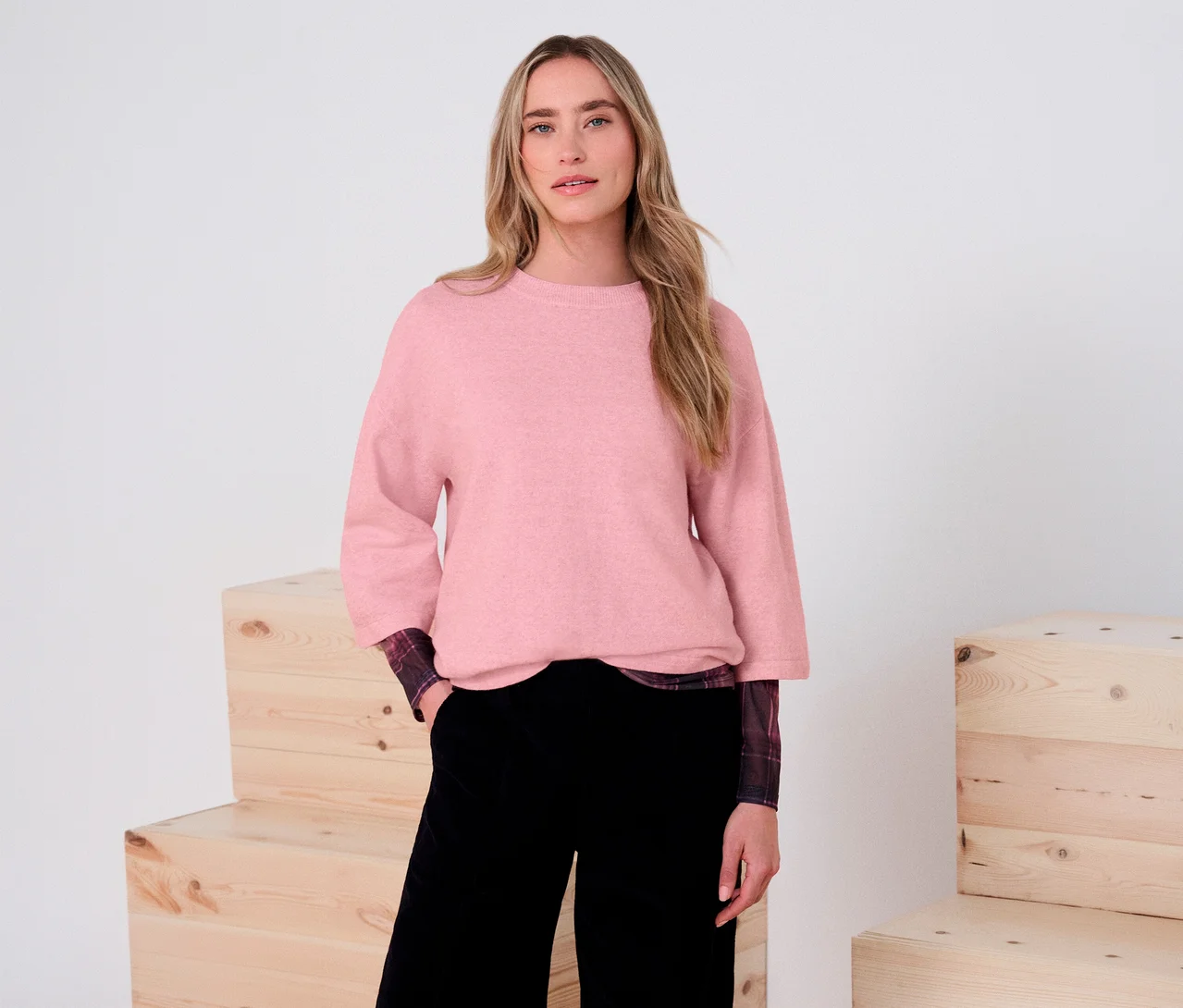 Eine Frau trägt einen rosa Street One 3/4-Strickpullover, ein kariertes Hemd und schwarze Hosen. Sie steht vor einem weißen Hintergrund mit Holzkisten.