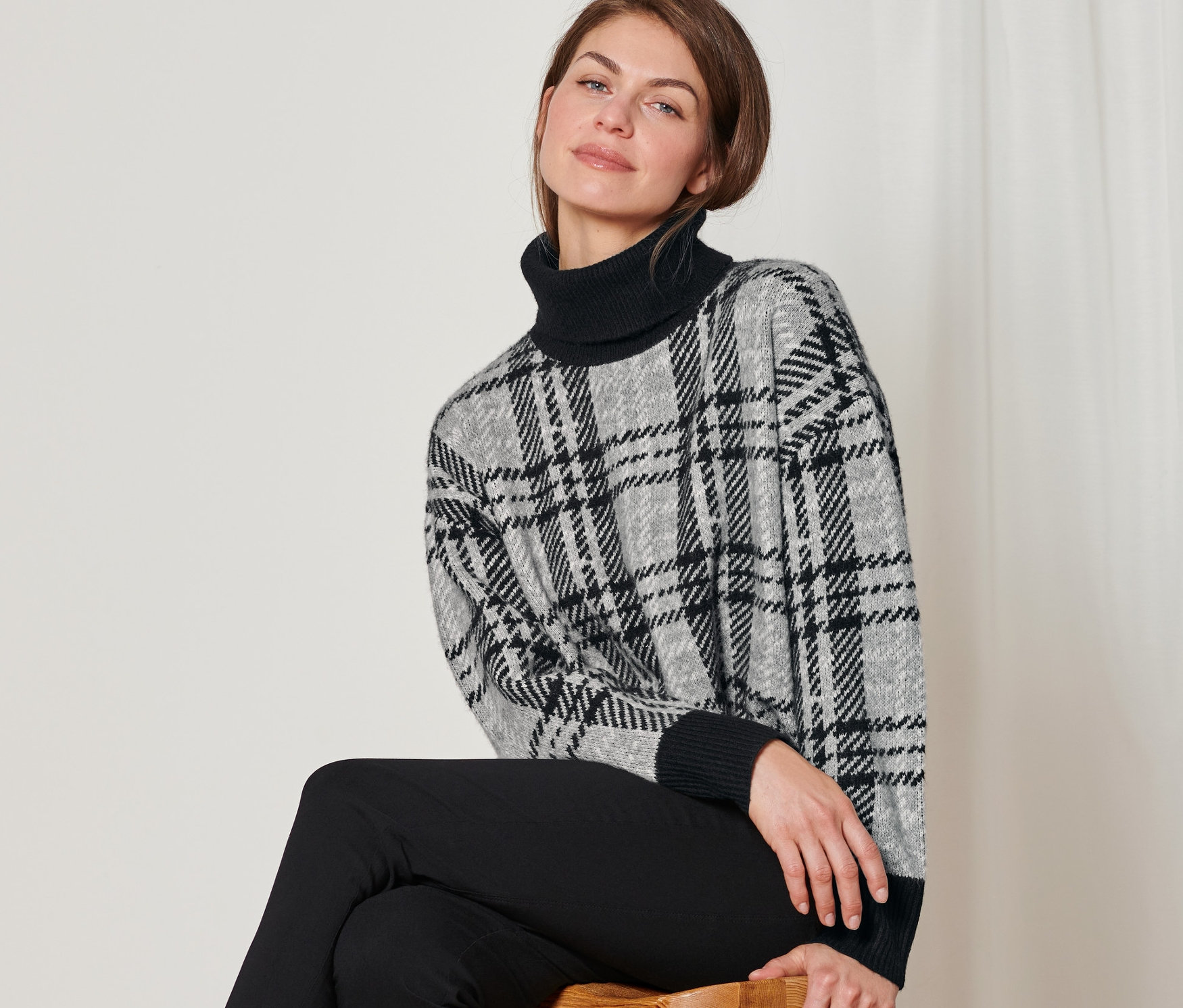 Frau sitzt auf einem Hocker und trägt einen Strickpullover mit Rollkragen.