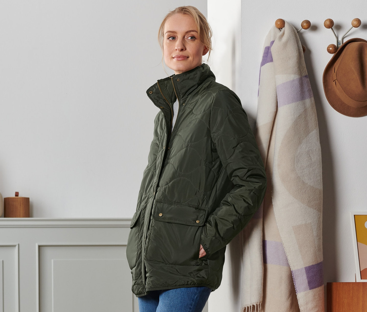 Frau lehnt an Wand und trägt Steppjacke und Jeans. Im Hintergrund Garderobe, Decke und Hut.