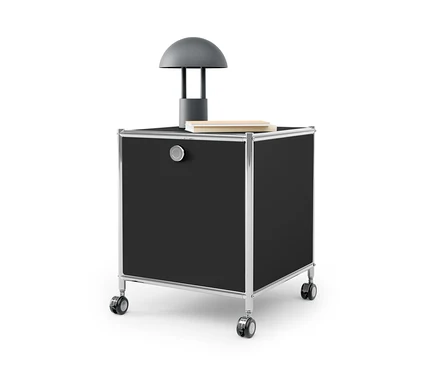 Ein schwarzer Beistelltisch mit Schublade Metall »CN3« mit silbernem Rahmen und Rädern. Auf dem Tisch stehen eine graue Lampe und ein Buch.