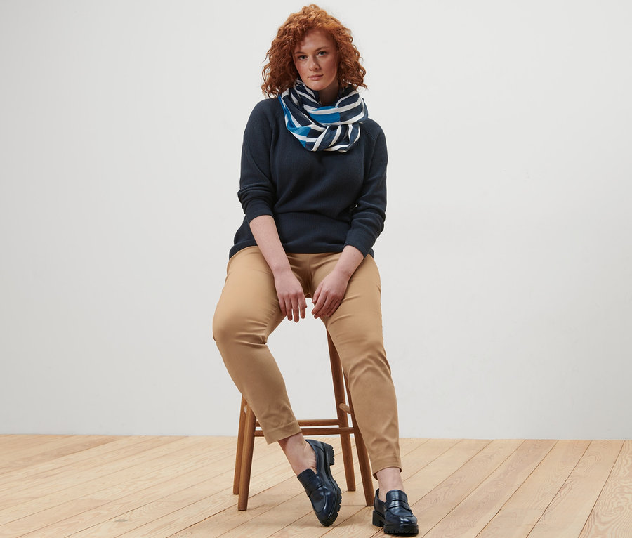 Frau sitzt auf einem Hocker und trägt einen dunkelblauen Strickpullover, einen gestreiften Loop-Schal und eine Stretchhose.