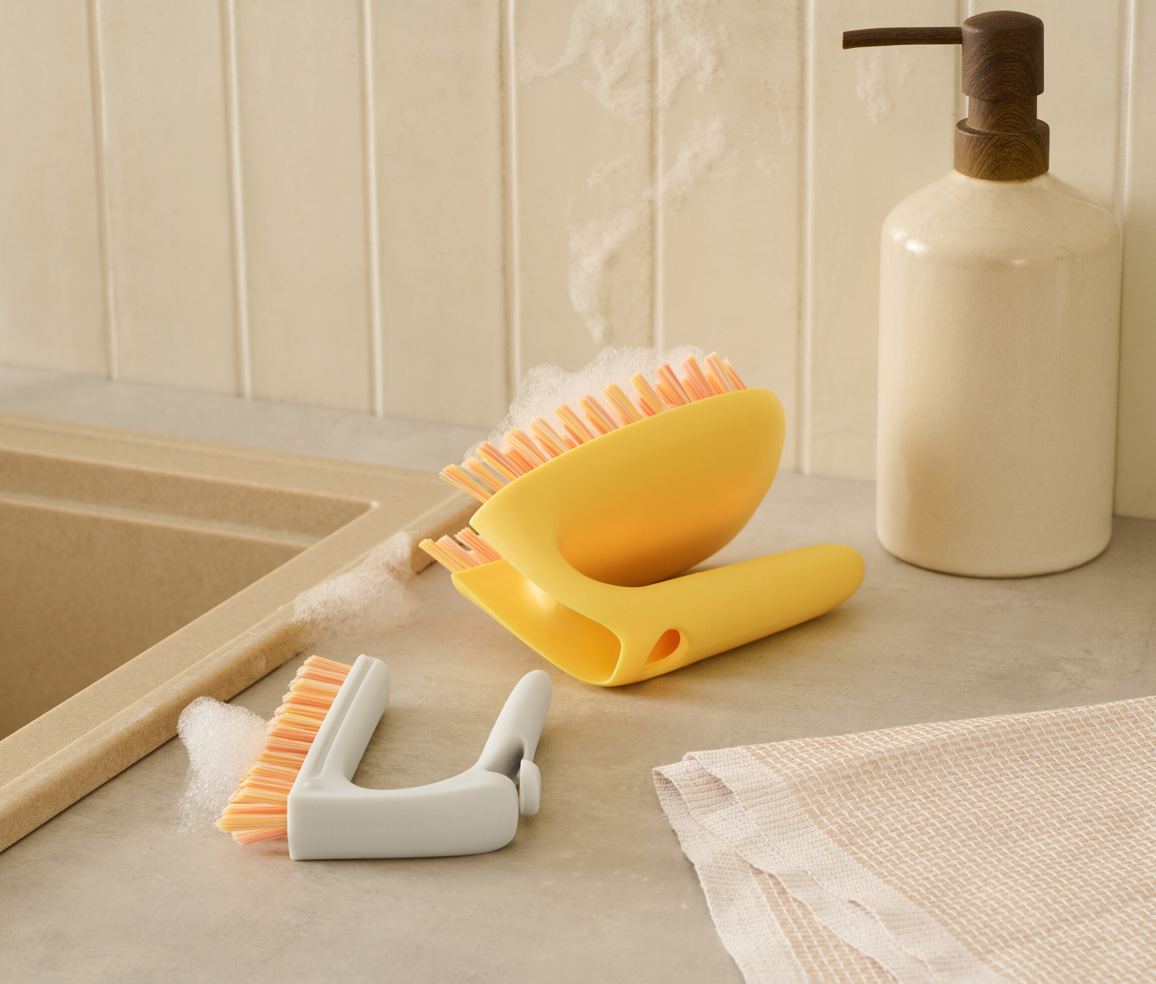 Eine gelbe 2-in-1-Fliesen-Bodenbürste und eine weiße Detailbürste liegen nebeneinander auf einer Theke neben einem Spülbecken.