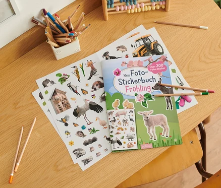 Auf einem Tisch liegen ein offenes Buch "Mein Foto-Stickerbuch – Frühling", ein Bogen mit Aufklebern, Buntstifte und ein Abakus.