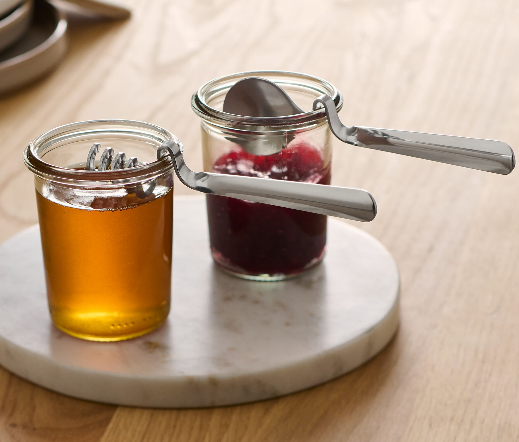 Zwei Gläser mit Honig und Marmelade stehen auf einem Marmortablett, in jedem Glas ein Marmeladen- und Honiglöffel.