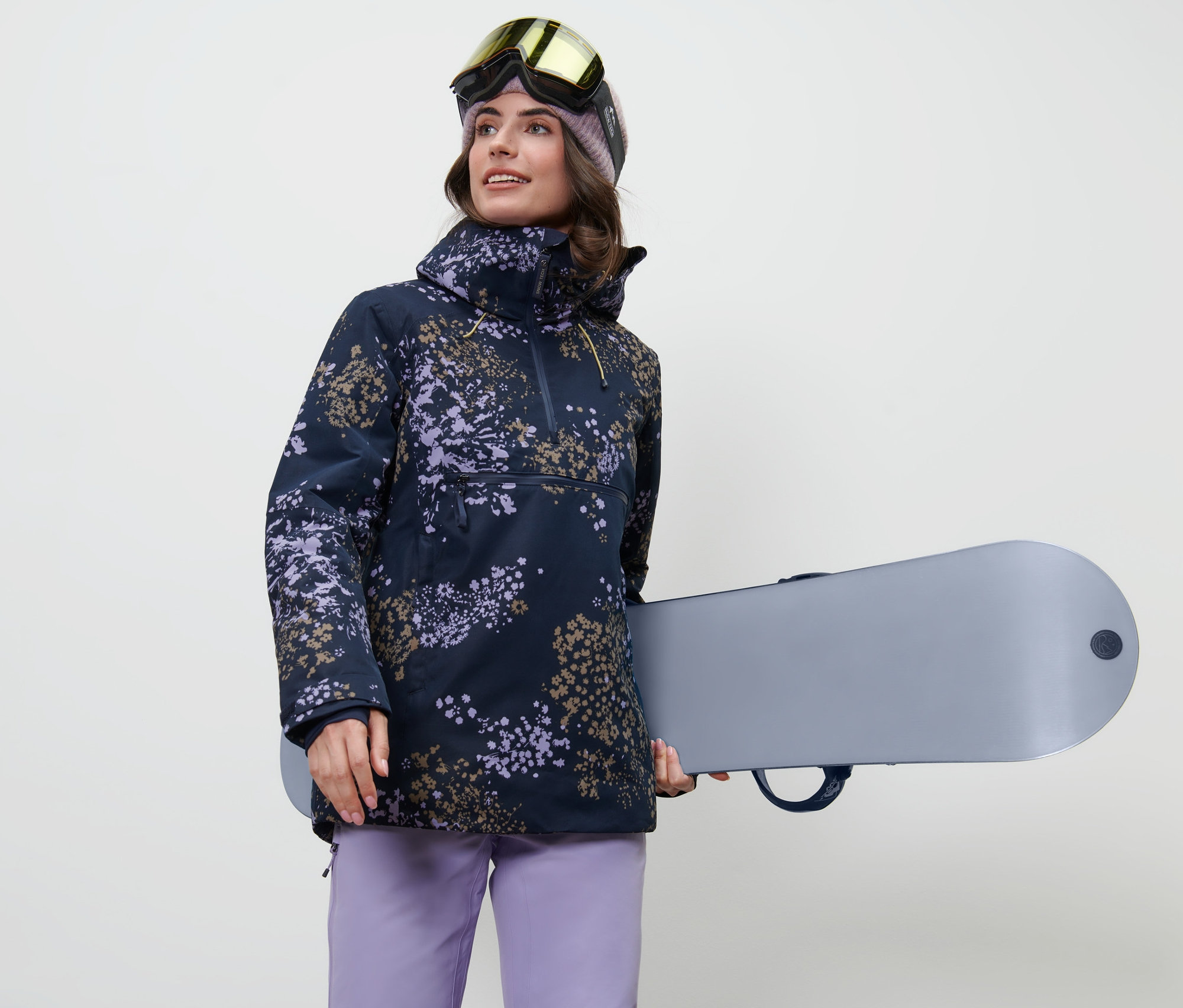 Frau hält ein Snowboard und trägt eine Halfzip-Skijacke.
