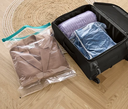 Ein beiger Mantel in einem Vakuum-Reisebeutel, neben einem schwarzen Koffer mit einem lila Pullover und einer Jeansjacke in Vakuum-Reisebeuteln.