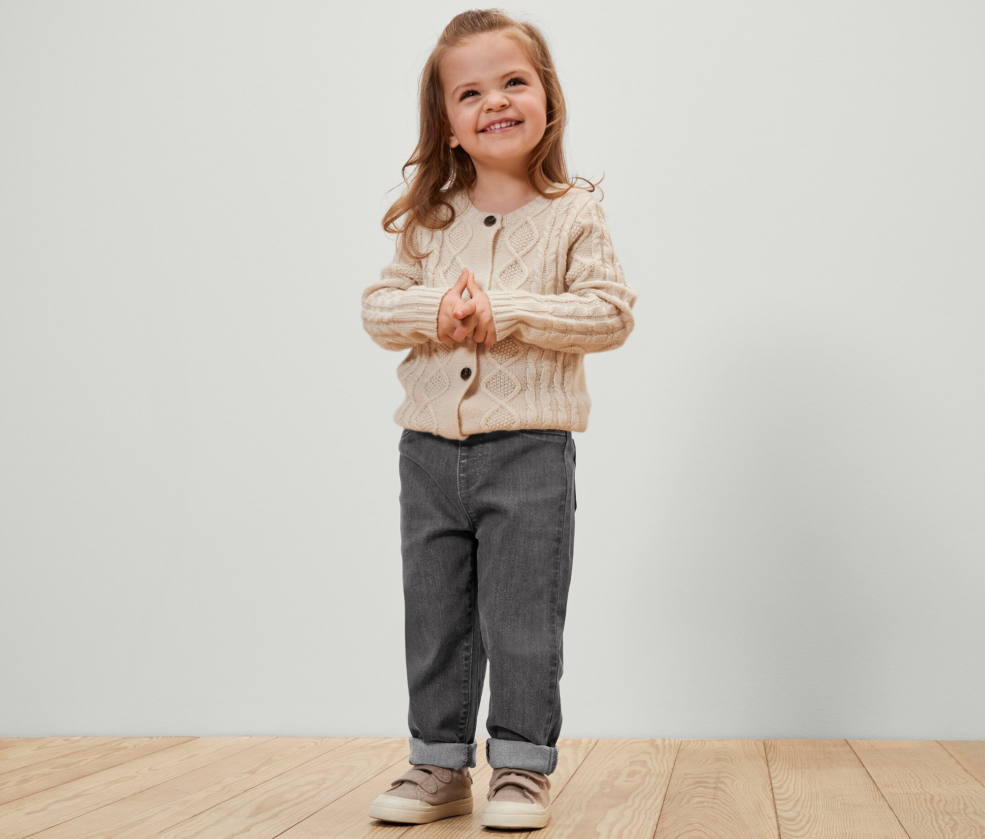 Lächelndes kleines Mädchen steht mit gefalteten Händen da. Sie trägt eine graue Hose und eine cremefarbene Strickjacke. Kinder-Pull-on-Pants – Fit »Luca«, grau.
