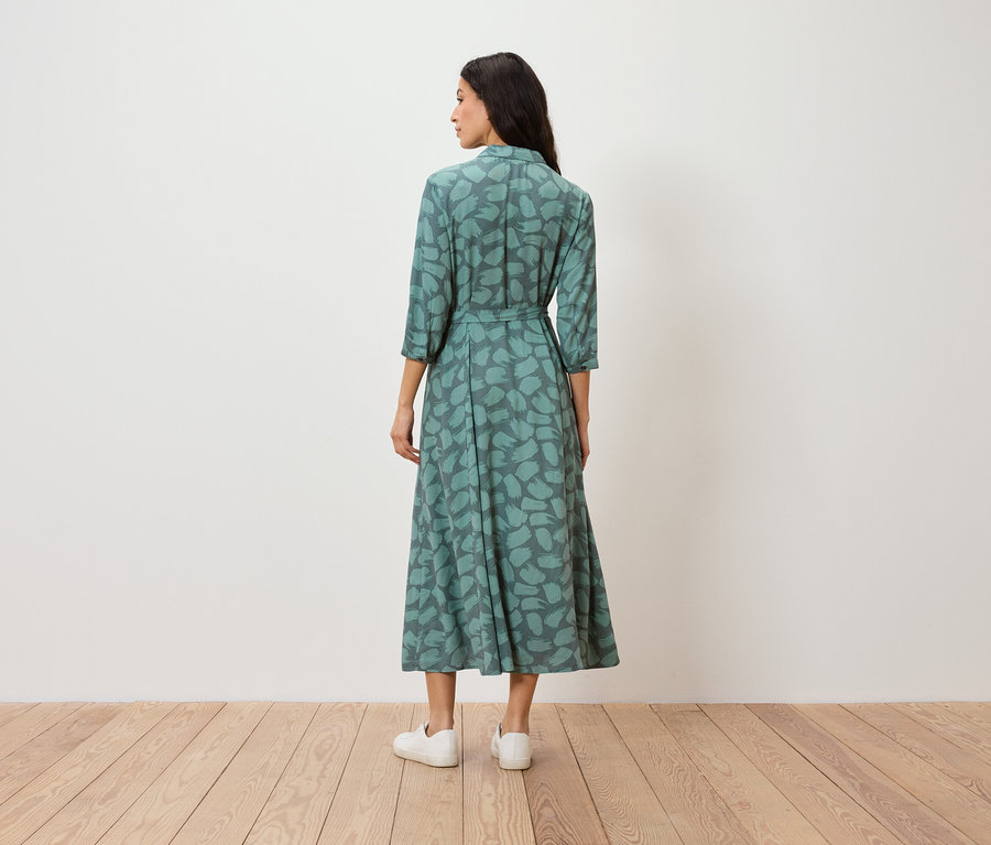 Eine Frau steht mit dem Rücken zur Kamera und trägt ein grünes, gemustertes Kleid und weiße Turnschuhe.