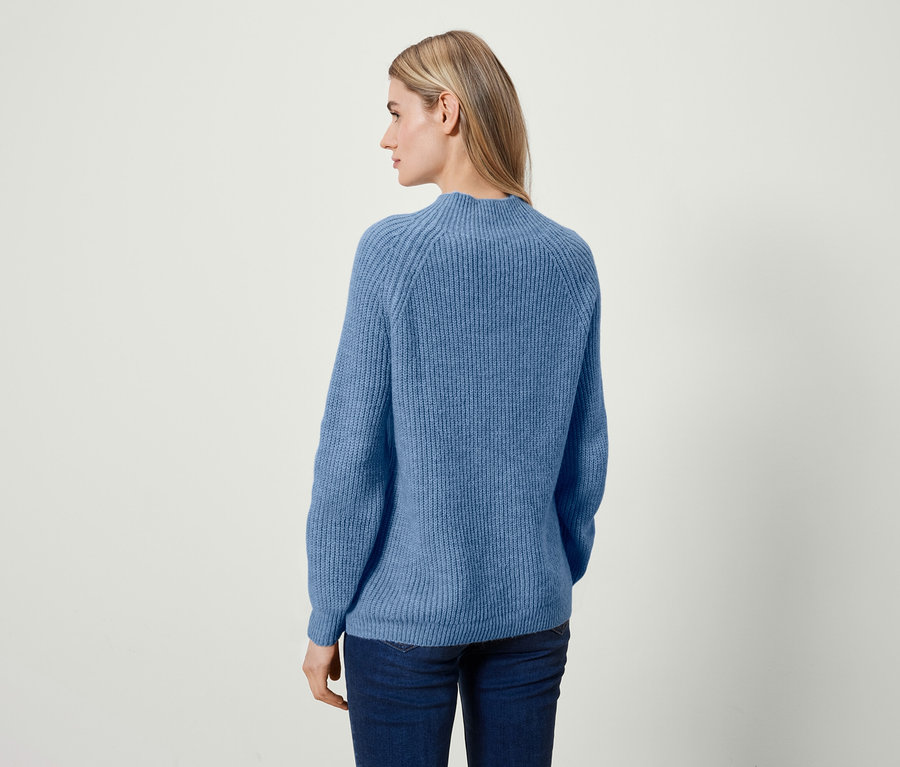 Eine Frau mit blonden Haaren steht mit dem Rücken zur Kamera und trägt einen blauen Pullover und Jeans.