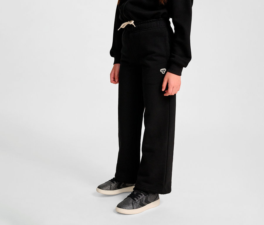 Nahaufnahme einer Person, die schwarze HUMMEL HMLJR Loose WL Sweatpants und Turnschuhe vor einem grauen Hintergrund trägt.