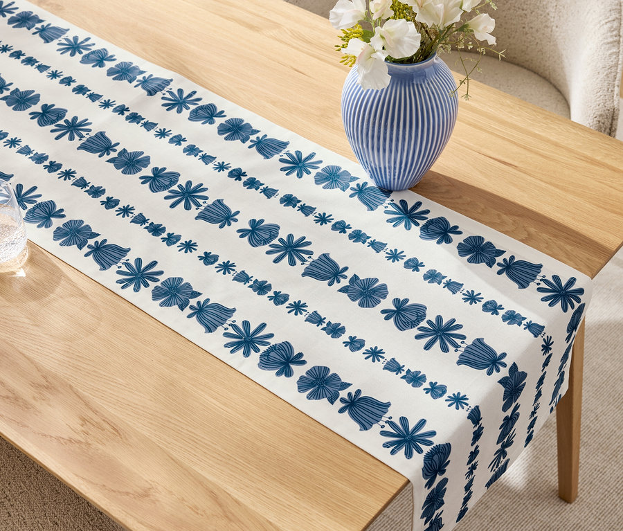 Beiger Holztisch mit blau-weissem Tischläufer, blauer Vase mit weißen Blumen und Glas.