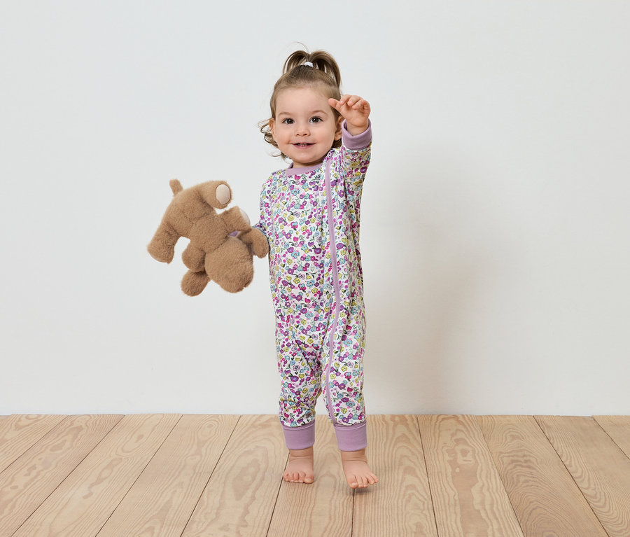 Ein Kind in einem geblümten Overall steht mit einem Plüschteddybär.