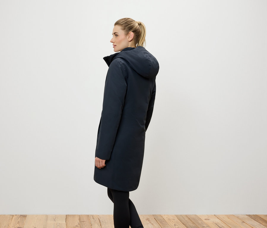 Seitenansicht einer Frau mit blonden, zum Pferdeschwanz zusammengebundenen Haaren, die einen langen, dunkelblauen Mantel mit Kapuze und schwarze Leggings trägt.