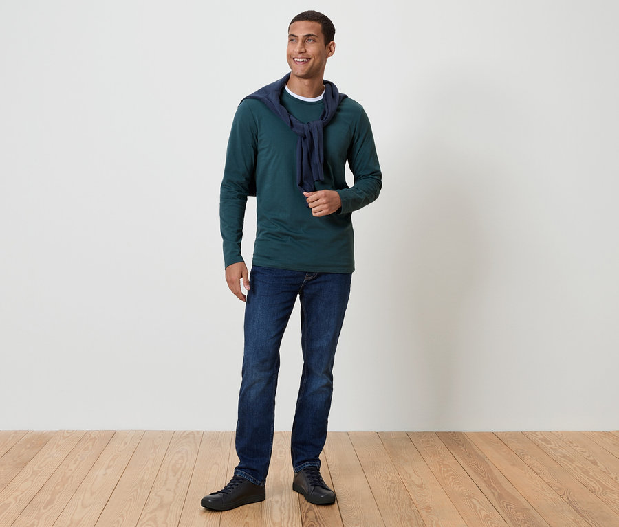 Ein Mann mit dunklem Haar und einem Lächeln steht auf einem Holzboden. Er trägt ein grünes Langarmshirt, blaue Jeans und schwarze Schuhe. Um seinen Hals ist ein dunkelblauer Pullover drapiert.