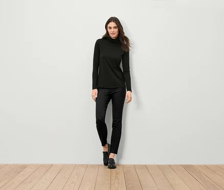 Eine Frau steht und trägt einen schwarzen Rollkragenpullover, eine schwarze Hose und schwarze Schuhe.