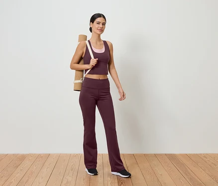 Frau in Sportbekleidung mit Yogamatte über der Schulter steht auf Holzboden vor einer weißen Wand.