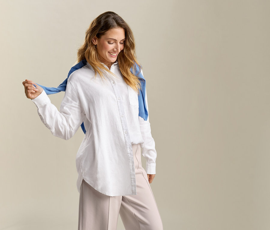 Frau mit blauem Pullover über den Schultern trägt eine lange, weiße Leinenbluse.