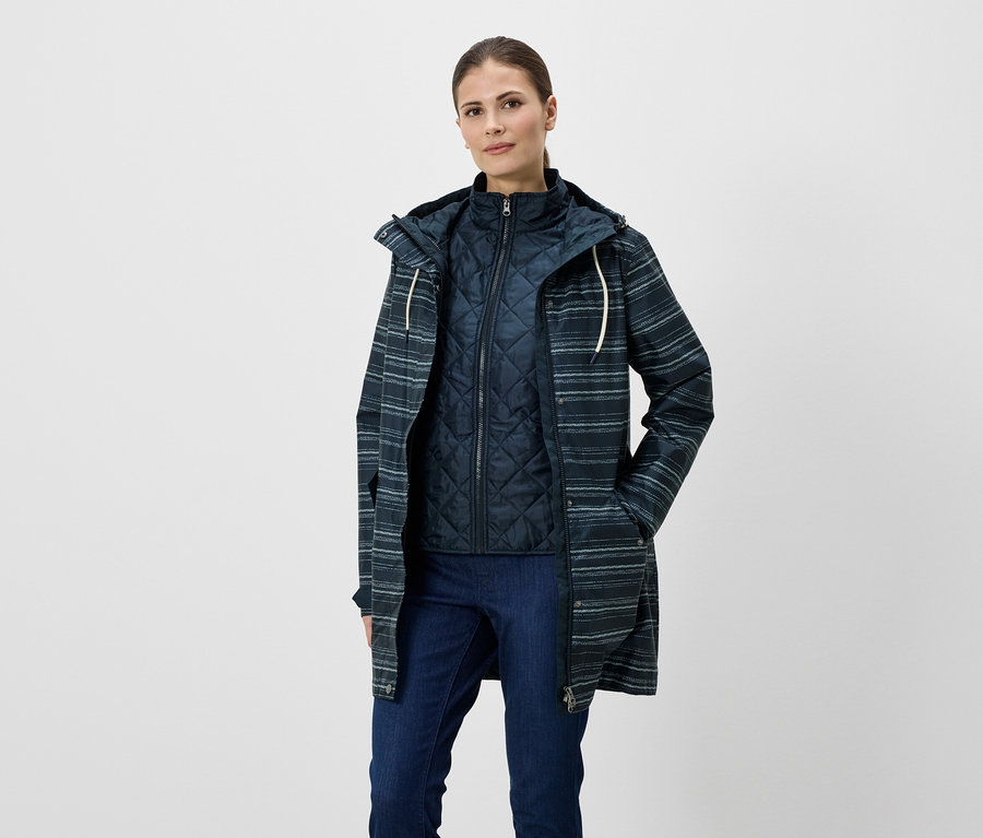 Frau mit Jeans, Steppweste und gestreifter Regenjacke mit Kapuze. Die Jacke hat Taschen und eine helle Kordel.