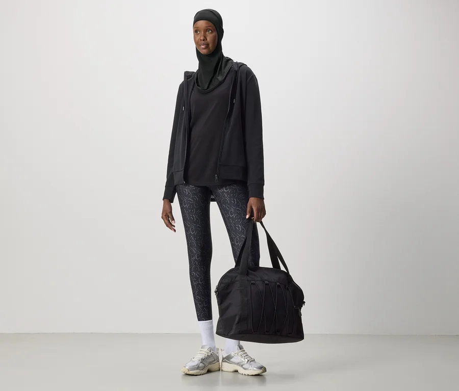 Frau mit schwarzem Hijab, Kapuzenjacke, Leggings mit Muster und Turnschuhen hält eine schwarze Sporttasche.