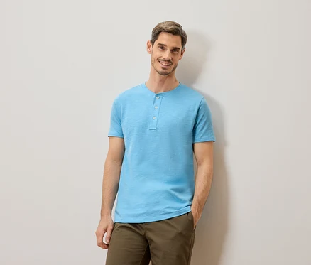 Ein Mann mit kurzen braunen Haaren und Bart, der ein hellblaues T-Shirt mit Knöpfen und eine Khakihose trägt, steht vor einer hellgrauen Wand.