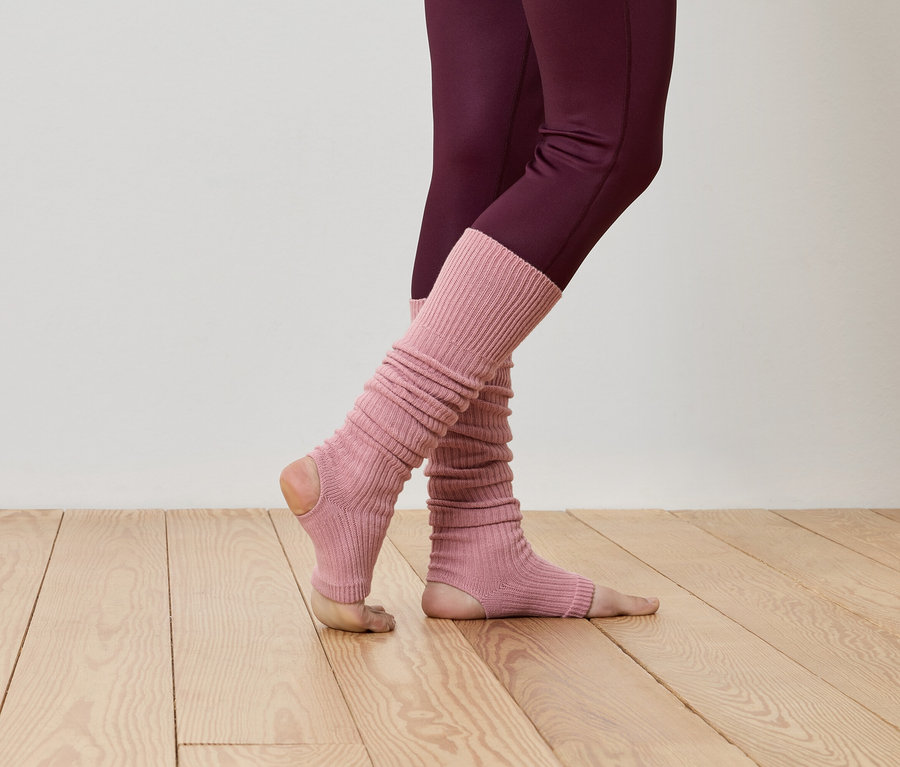 Nahaufnahme einer Person in bordeauxfarbenen Leggings und gerippten, rosafarbenen Stulpen auf einem Holzboden.