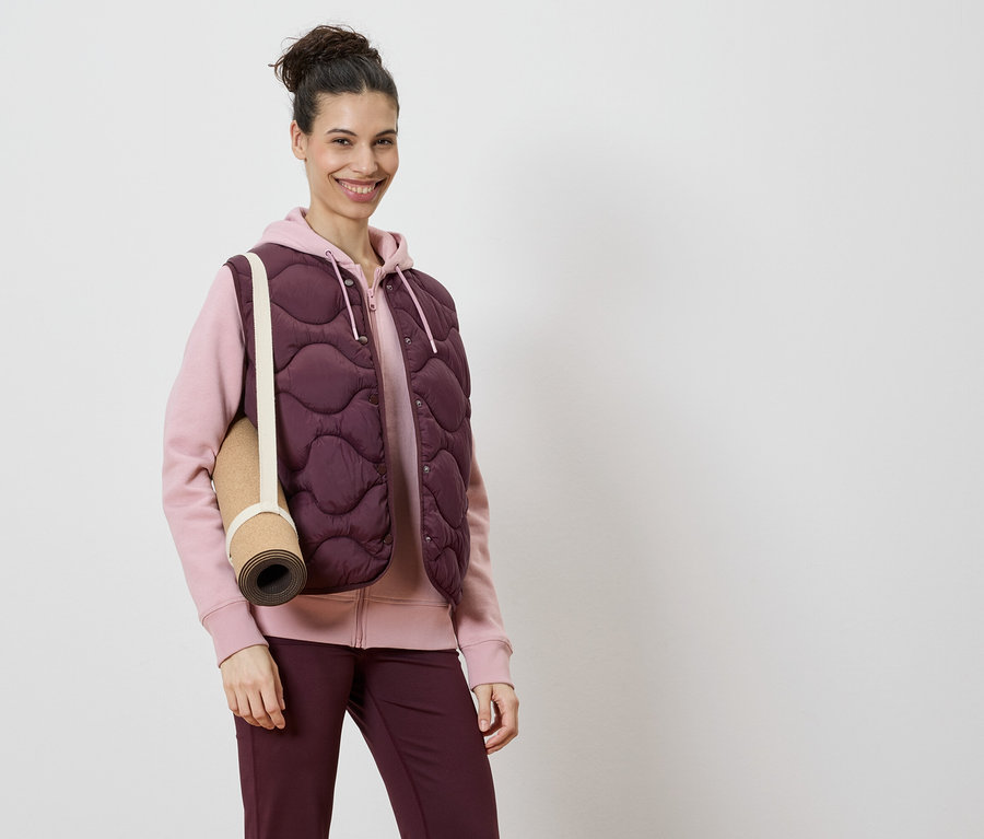 Frau trägt eine burgunderfarbene Steppweste über einem rosa Kapuzenpullover und burgunderfarbene Leggings, mit einer Yogamatte über der Schulter.