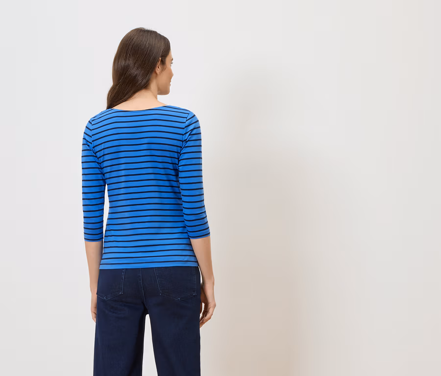 Rückansicht einer Frau mit braunen Haaren in einem blau-schwarz gestreiften Oberteil und dunklen Jeans vor einem weißen Hintergrund.