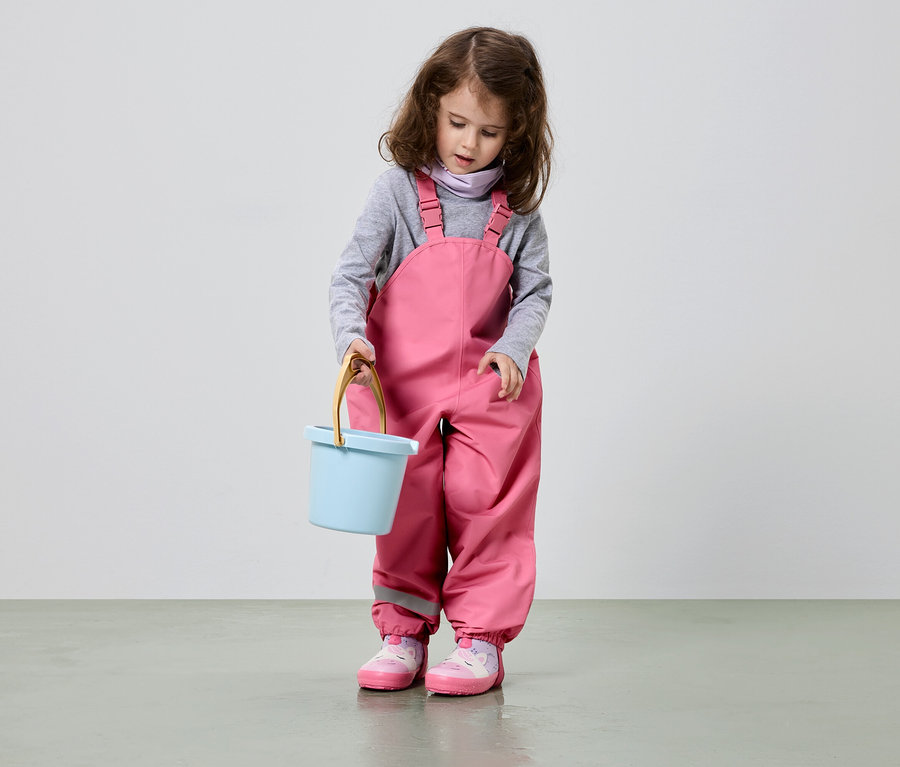 Ein Mädchen mit braunen Locken hält einen hellblauen Eimer und trägt eine rosa Latzhose und rosa Schuhe.