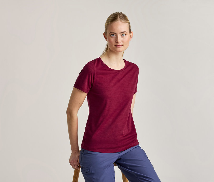 Eine Frau mit zurückgebundenen blonden Haaren, die ein weinrotes Kurzarm-T-Shirt und blaue Hosen trägt, sitzt auf einem Holzschemel und blickt in die Kamera.