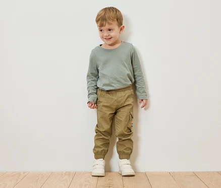 Ein Junge mit blonden Haaren steht vor einer weißen Wand und trägt ein grünes Langarmshirt, Khaki-Hosen und weiße Schuhe.
