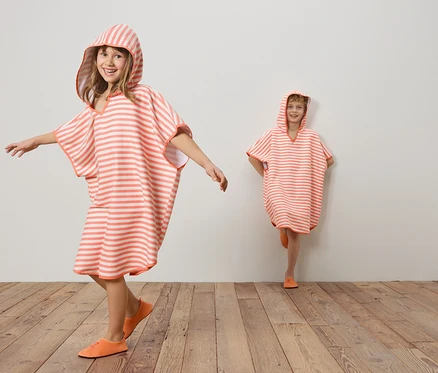 Zwei Kinder tragen gestreifte, korallenrote und weiße Kapuzenponchos und korallenrote Schuhe auf einem Holzboden vor einer weißen Wand.