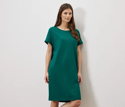 Frau trägt ein grünes, kurzes Kleid vor einer weißen Wand.
