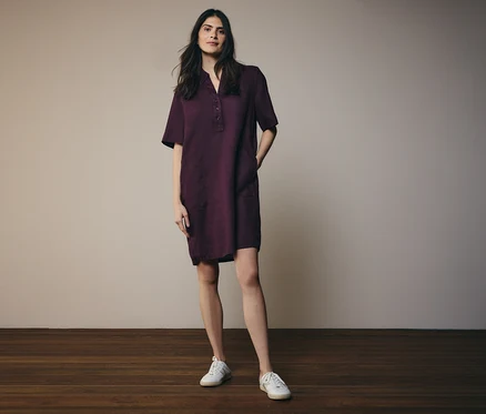 Frau in einem burgunderfarbenen Hemdkleid mit kurzen Ärmeln und weißen Turnschuhen, stehend auf einem Holzboden.