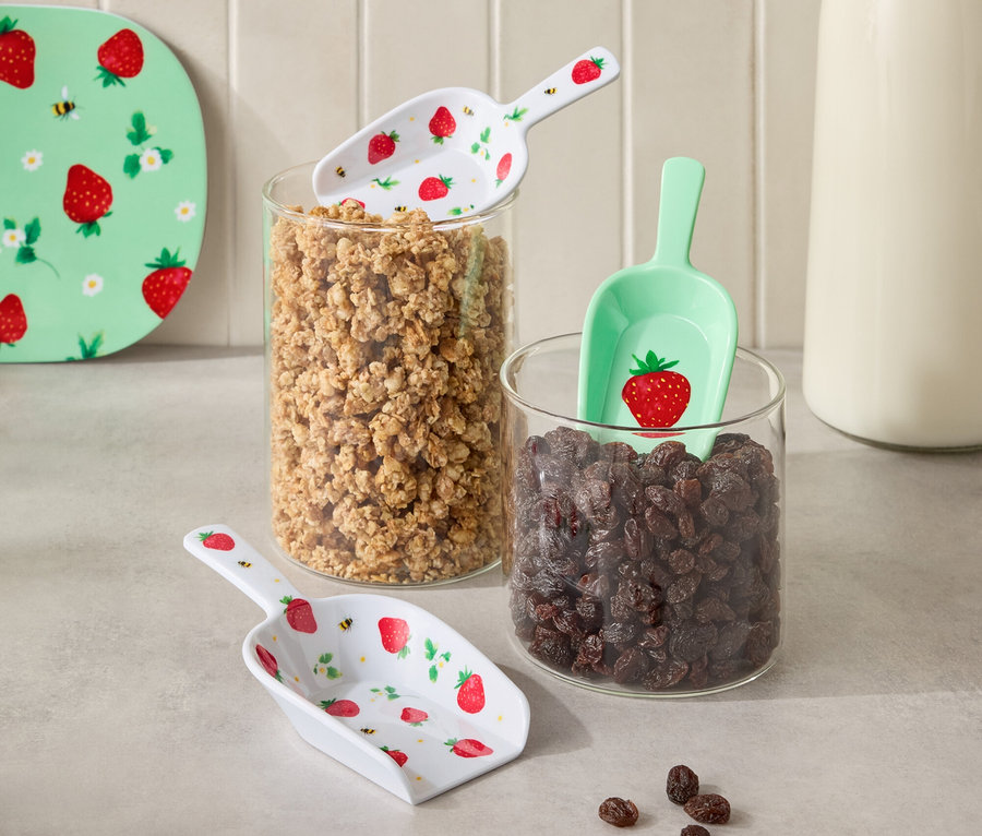 Abbildung von zwei Glasbehältern, gefüllt mit Müsli und Rosinen, dazwischen eine Flasche Milch. Auf den Behältern liegen Schaufeln mit Erdbeermuster.