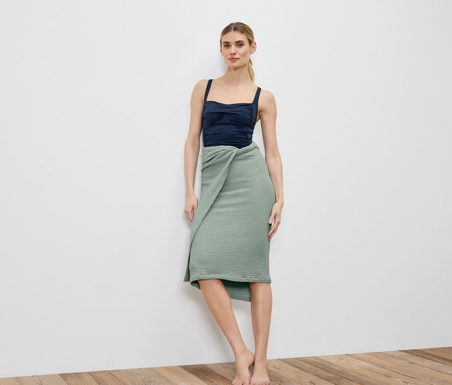 Frau steht barfuß vor einer weißen Wand, trägt ein marineblaues Top und einen gewickelten, hellgrünen Rock.