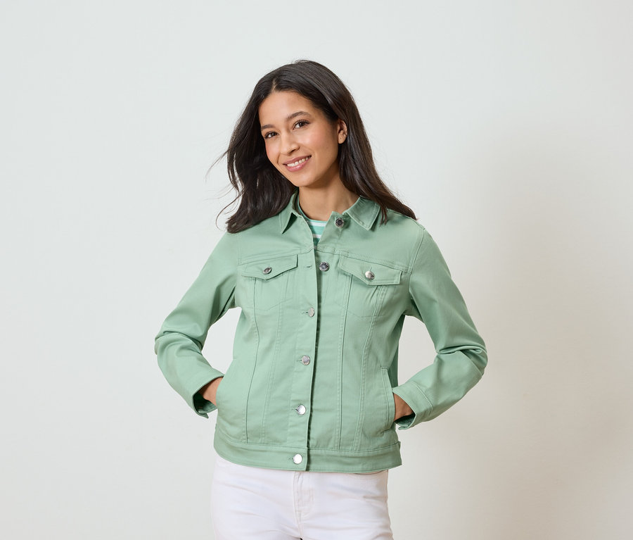 Frau mit hellgrüner Jeansjacke und weißen Hosen, die mit den Händen in den Taschen posiert.