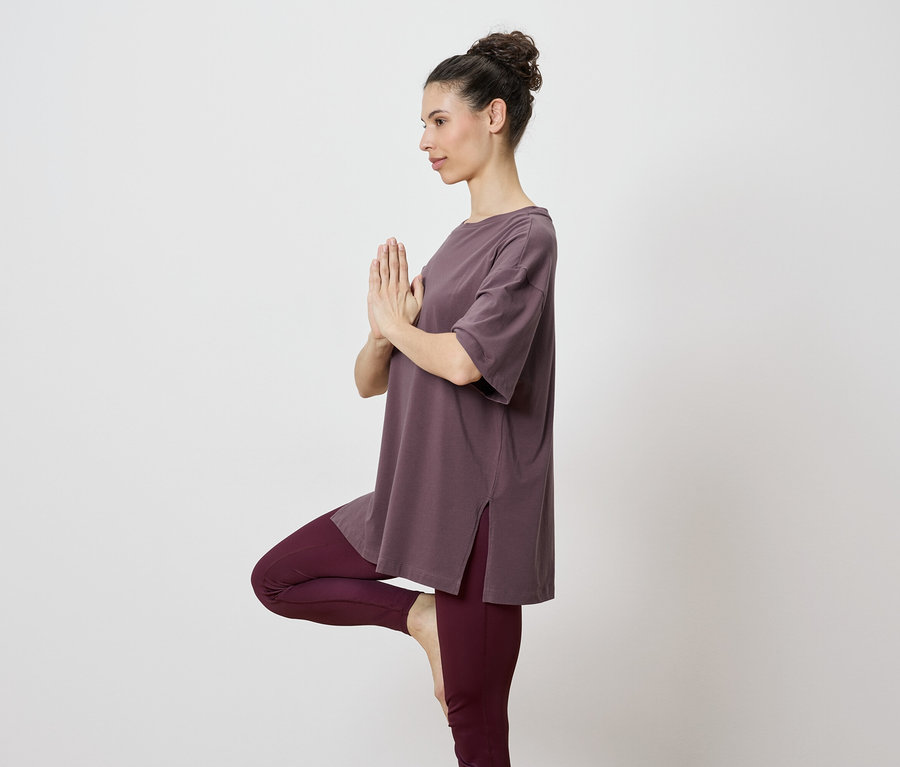 Frau in braunem Oberteil und Leggings, die eine Yoga-Pose auf einem Bein ausführt.