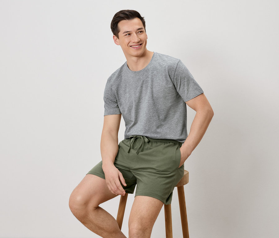 Mann sitzt auf einem Hocker, trägt ein graues T-Shirt und grüne Shorts.