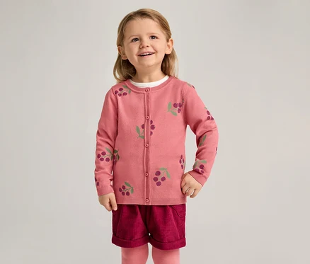Ein lächelndes Mädchen trägt eine rosa Strickjacke mit Kirschmuster, bordeauxrote Shorts und rosa Strumpfhosen vor einem hellen Hintergrund.