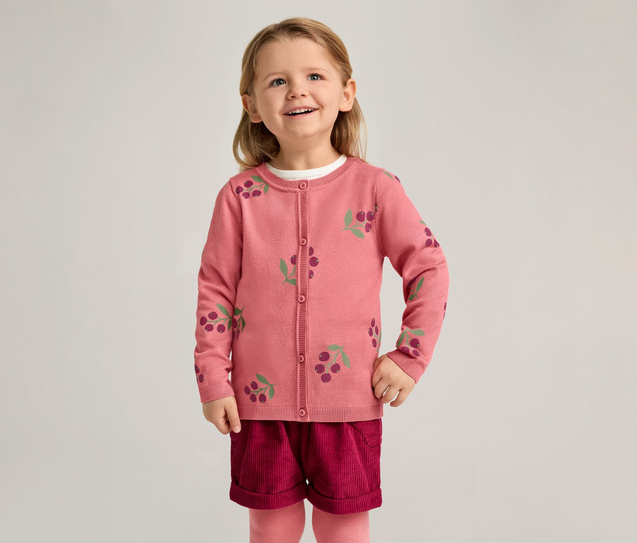 Ein lächelndes Mädchen trägt eine rosa Strickjacke mit Kirschmuster, bordeauxrote Shorts und rosa Strumpfhosen vor einem hellen Hintergrund.