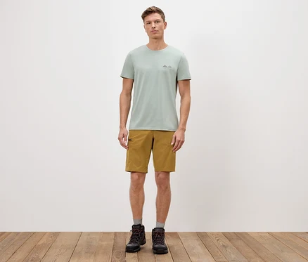 Mann steht vor einer weißen Wand und trägt ein grünes T-Shirt, Shorts und Stiefel.