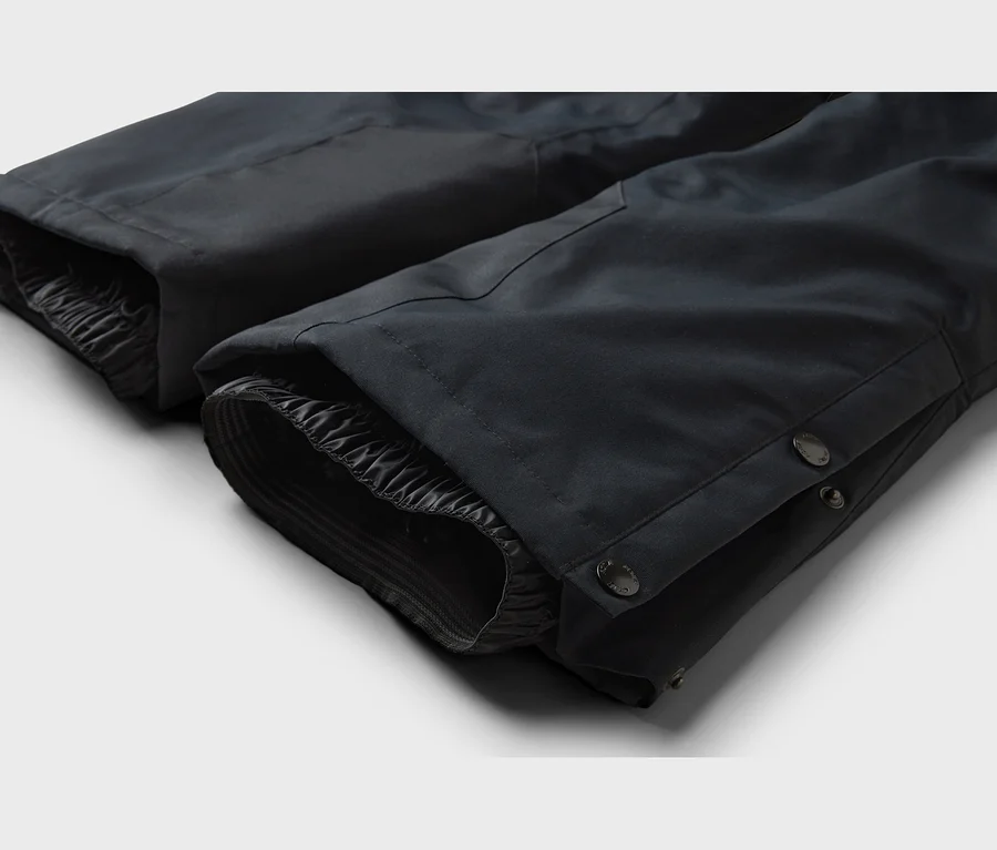 Nahaufnahme der unteren Beinabschlüsse einer dunklen Hose, mit elastischen Innenbündchen und Druckknöpfen am äußeren Saum.