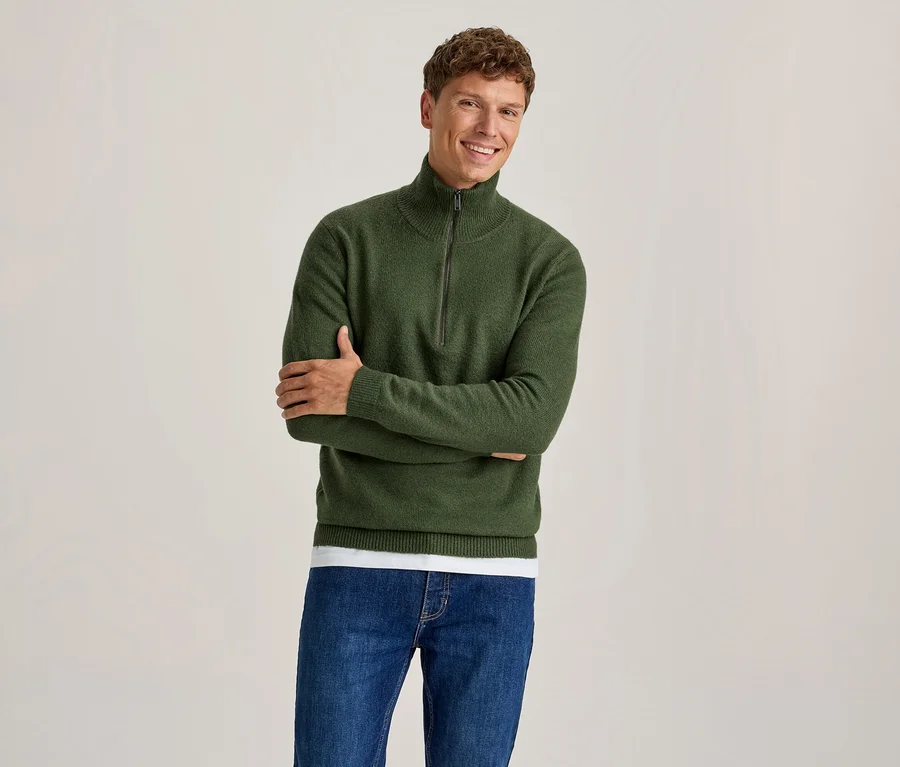 Lächelnder Mann in grünem Half-Zip-Pullover über einem weißen T-Shirt und blauen Jeans, die Arme verschränkt.