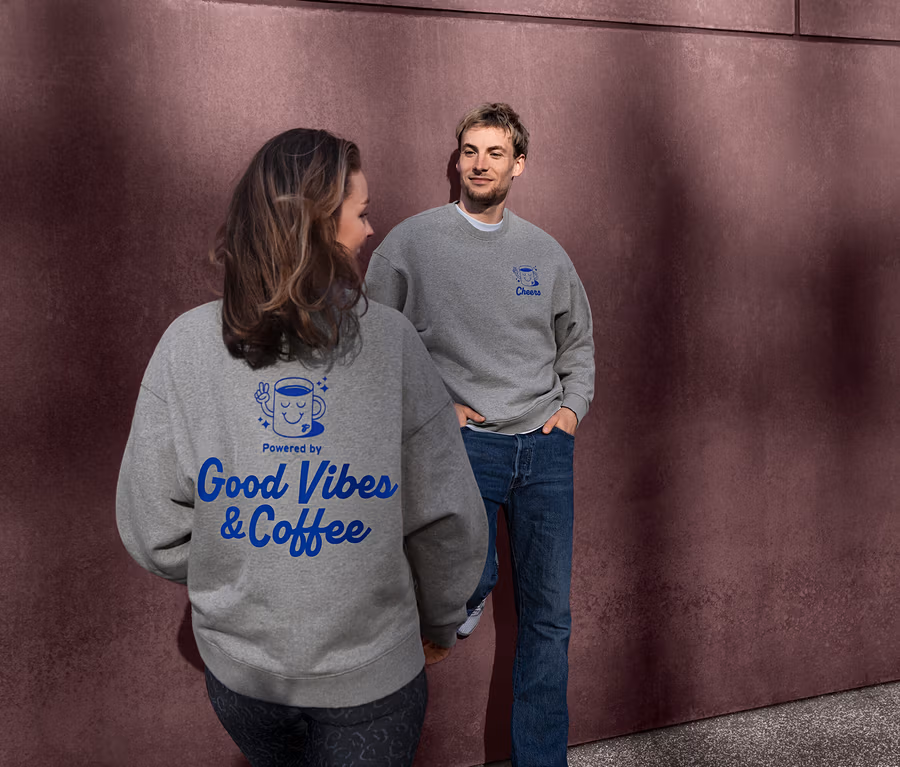 Frau und Mann tragen graue Unisex Coffee Crew Sweatshirts. Der Rücken des Sweatshirts der Frau zeigt ein blaues "Powered by Good Vibes & Coffee" Design. Der Mann blickt die Frau an, sein Sweatshirt hat ein blaues "Cheers" Design.