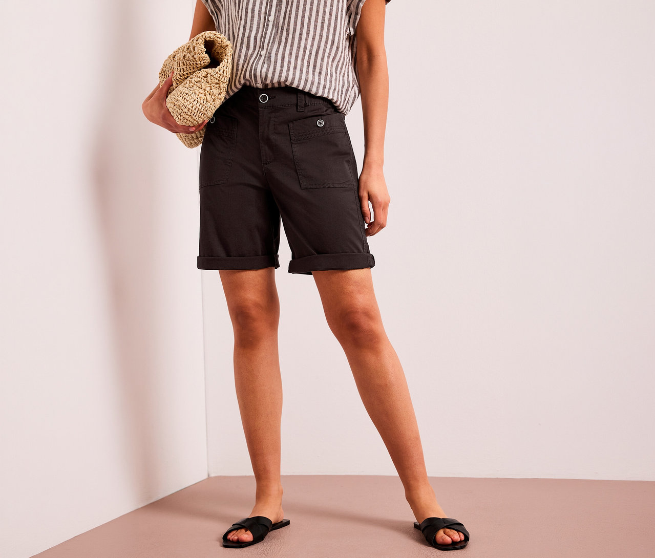 Eine Person trägt braune Street One Bermuda »Yulius« mit umgekrempelten Säumen, ein gestreiftes Oberteil und schwarze Sandalen und hält eine gewebte Tasche.