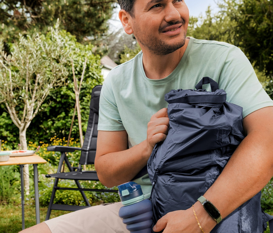Ein Mann in mintgrünem Outdoor-T-Shirt und beiger Shorts sitzt im Garten und hält einen dunkelblauen wasserdichten Rucksack und eine blaue faltbare Trinkflasche.
